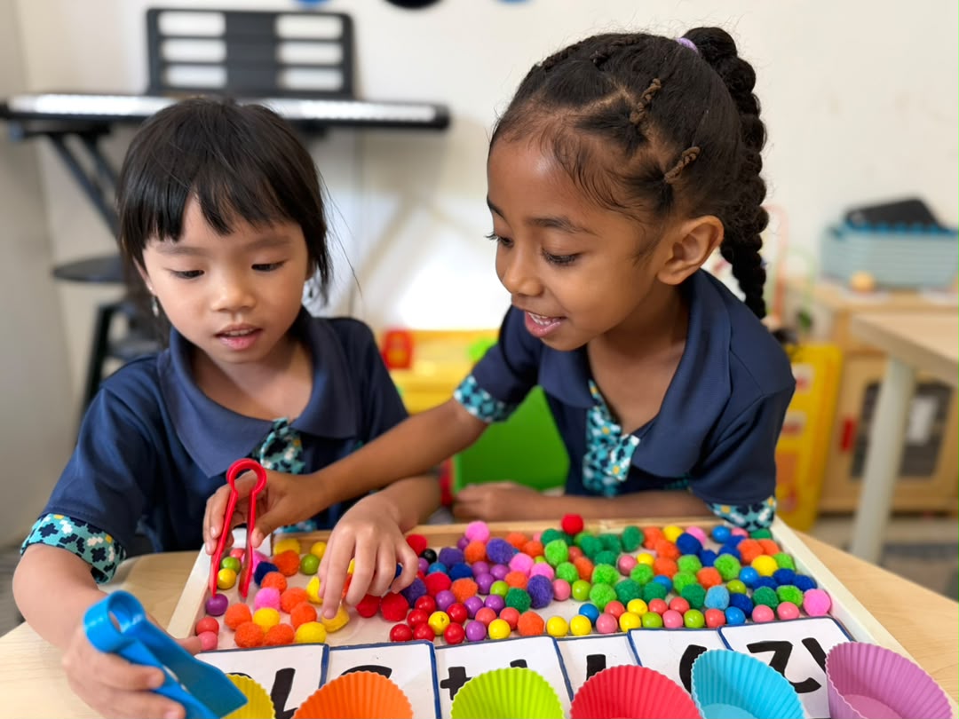 Two Early Years girls using tongs to pick up colourful pom-poms